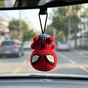 May include: A small, round, crocheted hanging ornament featuring a red and blue color scheme with a black web pattern. The object has two white, almond-shaped eye patches outlined in black, resembling a superhero mask, and hangs from a black cord.