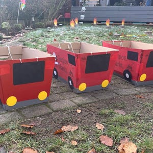 Könnte beinhalten: Drei rote Pappkartons in Form von Feuerwehrautos, mit schwarzen Fenstern, gelben Scheinwerfern und grauen Stoßfängern. Die Kartons stehen auf einer Ziegeloberfläche mit abgefallenen Blättern.