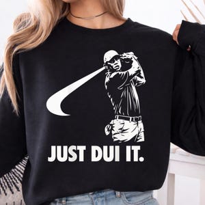 May include: Black sweatshirt with a white graphic of a golfer swinging a club, with a Nike swoosh. The text "JUST DUI IT." is printed in white below the graphic. The sweatshirt is a casual style.