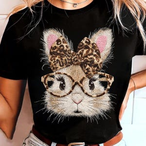 May include: Black t-shirt featuring a detailed illustration of a bunny wearing glasses and a leopard print bow. The bunny has pink inner ears and a light brown face. The glasses are a tortoise shell pattern.