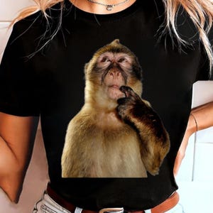 以下が含まれることがあります： 考え事をしているかのように、口に指を当てた猿のリアルなイメージがプリントされた黒いTシャツ。猿は茶色とタン色の毛皮をしています。シャツはクルーネックで半袖です。