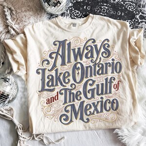 May include: A cream-colored short-sleeve t-shirt featuring decorative blue and red typography that reads Always Lake Ontario and The Gulf of Mexico, accented with intricate swirling line art patterns across the front of the garment.