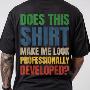Peut inclure: Un t-shirt noir avec un texte multicolore au dos qui indique : DOES THIS SHIRT MAKE ME LOOK PROFESSIONALLY DEVELOPED ? Les mots sont imprimés en vert, bleu, beige, jaune et rouge, avec une texture vieillie et un aspect vintage.