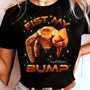 Può includere: T-shirt nera con le parole "FIST MY BUMP" in lettere arancioni e dorate. Il design presenta un pugno stilizzato e strutturato e un motivo a stelle. È incluso anche il testo "PROJECT HAIL MARY".