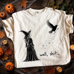 Op de afbeelding: Een crèmekleurig t-shirt met een zwarte afbeelding van een heks met een puntmuts die opkijkt naar een vliegende kraai met een toverstaf met ster. Onder de kraai daalt een spoor van sterren af naar de tekst "well, shit.." in het zwart.