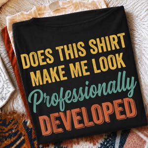 Puede incluir: Una camiseta negra con el texto amarillo "DOES THIS SHIRT MAKE ME LOOK", el texto cursivo verde azulado "Professionally" y el texto naranja "DEVELOPED". La camiseta está sobre una prenda de color crema y una prenda de color óxido.