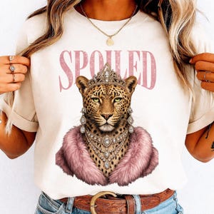 May include: Cream-colored t-shirt featuring a regal leopard illustration. The leopard wears a crown, jeweled earrings, and a pink fur stole. The word "SPOILED" is printed above the leopard in pink letters. A gold necklace is also visible.
