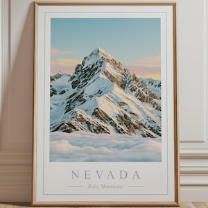 May include: A framed vertical wall art print featuring a snow-capped mountain peak rising above a layer of clouds. The sky displays soft pink and blue hues. The text NEVADA and Ruby Mountains is printed in a light blue font at the bottom of the print.