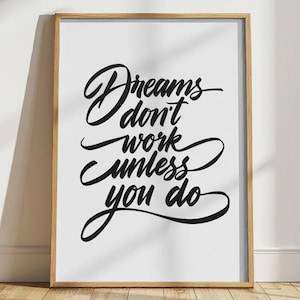 Peut inclure: Une affiche encadrée avec la citation "Dreams don't work unless you do" en écriture noire élégante sur fond blanc. Le cadre est de couleur marron clair, et l'affiche est posée sur un sol en bois.