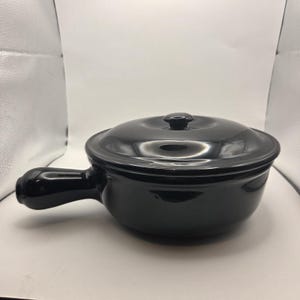 May include: A black ceramic soup bowl with a lid and a handle. The bowl is round with a slightly flared rim and a matching lid with a small knob. The handle is curved and attached to the side of the bowl. The bowl is designed for serving soups and stews.