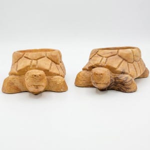 May include: Two carved wooden turtle-shaped bowls with visible wood grain patterns. Each turtle features a detailed shell design on the back, a rounded head, and four legs. The light brown wood has natural variations in color and texture throughout.