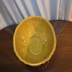 Op de afbeelding: Een ovale keramische kom in mosterdgeel met een gespikkelde afwerking. De rand heeft een geschulpte rand. De kom is leeg en staat op een donker houten oppervlak. De binnenkant van de kom is een donkerdere tint geel.