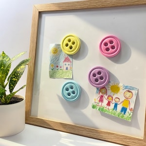 Puede incluir: Una pizarra enmarcada en madera con cuatro imanes grandes en forma de botón de colores brillantes. Dos dibujos infantiles están sujetos a la pizarra. A la izquierda hay una planta en maceta con hojas verdes y amarillas.