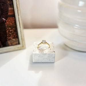 May include: A gold ring featuring a central oval-cut clear gemstone, set on a thin band. The ring is displayed on a small, square, white speckled stone block. The design highlights a classic metal setting with a single prominent stone.