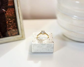 Puede incluir: Un anillo de oro con una piedra preciosa transparente de corte ovalado en el centro, montado sobre una banda fina. El anillo se exhibe sobre un pequeño bloque de piedra cuadrado de color blanco moteado. El diseño destaca un engaste clásico.