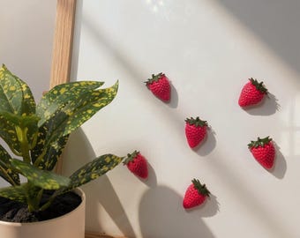 Op de afbeelding: Vijf kleine, felrode aardbeivormige magneten met groene blaadjes zijn gerangschikt op een wit magneetbord met een lichthouten frame. De magneten hebben een getextureerd oppervlak en zijn verspreid over het bord geplaatst.