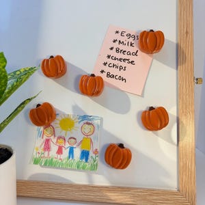 May include: A white magnetic dry-erase board with a light wood frame featuring five orange pumpkin-shaped magnets. A pink sticky note with a handwritten grocery list and a child's colorful drawing of a family are attached to the board.