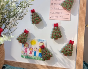 Op de afbeelding: Vijf groene kerstboommagneten met rode versieringen en een rode strik als piek, bevestigd aan een wit magneetbord. Het bord toont een roze papiertje met de handgeschreven tekst Grocery, Eggs, Bacon, Milk, Bread, cheese, en MUffins.