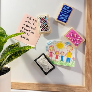 May include: A white magnetic board with a wooden frame features four rectangular pastry-shaped magnets, a pink note with handwritten text including Eggs, Milk, Bread, cheese, chips, and Bacon, and a child's drawing of a family under a yellow sun.