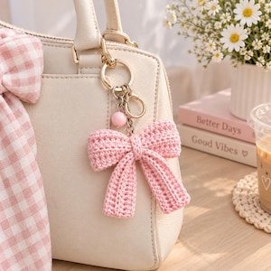 Può includere: Una borsa color crema con un portachiavi a forma di fiocco lavorato all'uncinetto in rosa, attaccato a un anello in metallo dorato. Il portachiavi include una piccola perla sferica rosa e un moschettone. Un fiocco in tessuto a quadretti rosa e bianco è legato al manico.