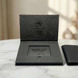 Könnte beinhalten: Ein schwarzes Portemonnaie und ein dunkelgrauer Kartenhalter mit dem Text "WEDJAT PROTECT YOURSELF" werden gezeigt. Der Kartenhalter hat ein Augensymbol und QR-Codes. Das Portemonnaie ist geschlossen und neben dem offenen Kartenhalter platziert.