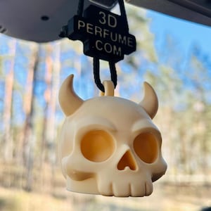 May include: A cream-coloured skull-shaped car air freshener with horns. The air freshener is suspended from a black tag that reads "3D PERFUME .COM". The skull has large eye sockets and a triangular nose cavity.