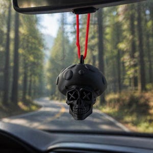 Puede incluir: Un ambientador de coche negro con forma de hongo y cara de calavera. Cuelga de un cordón rojo sujeto al espejo retrovisor de un coche. El fondo muestra una carretera a través de un bosque.