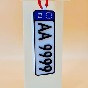 May include: A white rectangular license plate-shaped pendant with black text reading "AA 9999" and a blue EU emblem. The pendant hangs from a red cord attached to a black tag with "3D PERFUME .COM" printed on it.