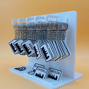 May include: A white display stand holding multiple square keychains with silver rings. Each keychain features a black and white graphic of a city skyline and text including names like Baltezers, Carnikava, Ādaži, Ventspils, and Sigulda.
