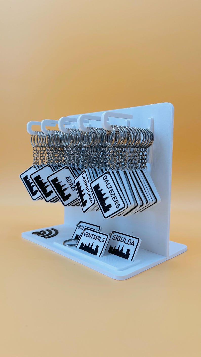 May include: A white display stand holding multiple square keychains with silver rings. Each keychain features a black and white graphic of a city skyline and text including names like Baltezers, Carnikava, Ādaži, Ventspils, and Sigulda.