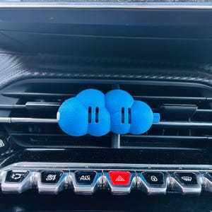 Può includere: Un profumatore per auto blu brillante a forma di nuvola, fissato a una bocchetta di ventilazione orizzontale. L'oggetto presenta una superficie opaca e testurizzata con due fessure verticali su ciascun lato della nuvola e una piccola linguetta.