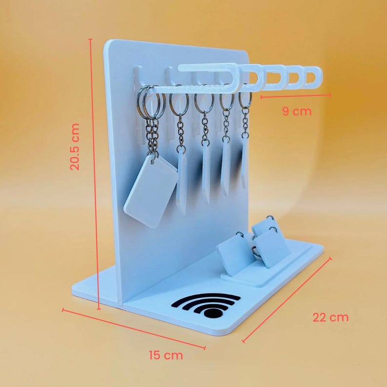 May include: A white display stand with a vertical panel featuring five hooks for keychains and a base with two slots for small rectangular signs. The stand measures 20.5 cm in height, 22 cm in width, and 15 cm in depth, with a 9 cm hook section.