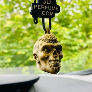 Könnte beinhalten: Ein olivgrüner, dreidimensionaler Lufterfrischer in Totenkopfform, der an einer schwarzen Kordel hängt. Über dem Totenkopf befindet sich ein schwarzes rechteckiges Schild mit dem weißen Text 3D PERFUME .COM. Der Totenkopf zeigt detaillierte Gesichtszüge, darunter eine Gehirnstruktur auf der Oberseite, tiefe Augenhöhlen und einen Kiefer mit entblößten Zähnen.