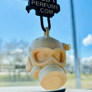 May include: Off-white, 3D-printed gas mask car air freshener. The mask has detailed features and is suspended by a black cord. The text "3D PERFUME .COM" is printed on a black tag above the mask. The background is a blue sky.