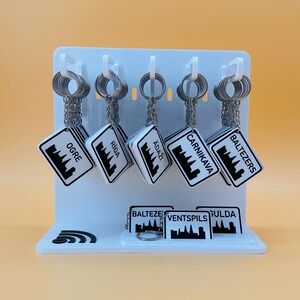 May include: A white display stand holding five hanging keychains and three additional keychains on the base. Each white square keychain features a black city skyline graphic and the text Ogre, Riga, Adaži, Carnikava, Baltezers, Ventspils, or Sigulda.