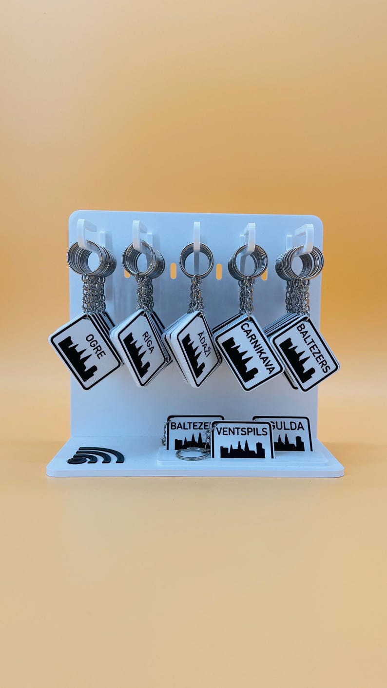 May include: A white display stand holding five hanging keychains and three additional keychains on the base. Each white square keychain features a black city skyline graphic and the text Ogre, Riga, Adaži, Carnikava, Baltezers, Ventspils, or Sigulda.