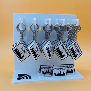 May include: A white display stand holding multiple square keychains with silver metal rings and chains. Each keychain features a black and white graphic of a city skyline and the text Ogre, Riga, Adazi, Carnikava, Baltezers, Ventspils, or Sigulda.