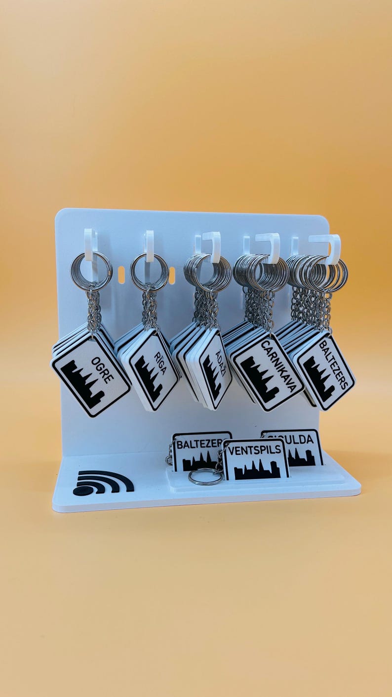 May include: A white display stand holding multiple square keychains with silver metal rings and chains. Each keychain features a black and white graphic of a city skyline and the text Ogre, Riga, Adazi, Carnikava, Baltezers, Ventspils, or Sigulda.