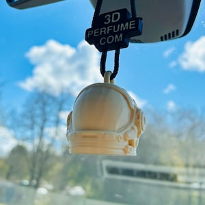 Könnte beinhalten: Ein cremefarbener Astronautenhelm-Auto-Lufterfrischer, der an einem Rückspiegel hängt. Der Lufterfrischer hängt an einem schwarzen Anhänger mit dem Text "3D PERFUME .COM". Der Hintergrund zeigt einen blauen Himmel mit Wolken.