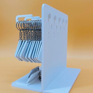 May include: A white vertical display stand with a flat base, featuring multiple rows of small holes for hanging items. A collection of silver metal key rings with attached white rectangular tags hangs from the top section of the display stand.