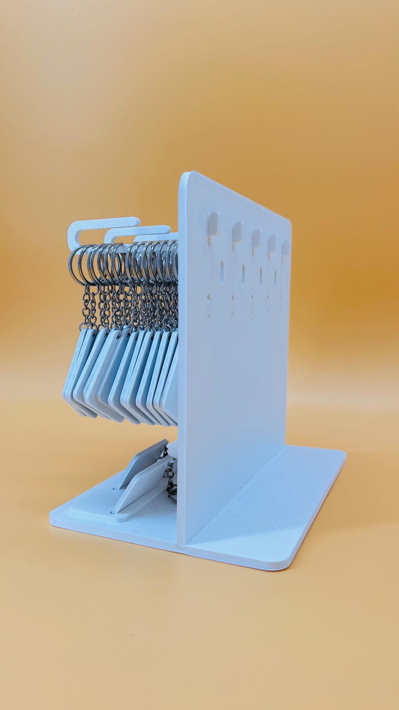 May include: A white vertical display stand with a flat base, featuring multiple rows of small holes for hanging items. A collection of silver metal key rings with attached white rectangular tags hangs from the top section of the display stand.