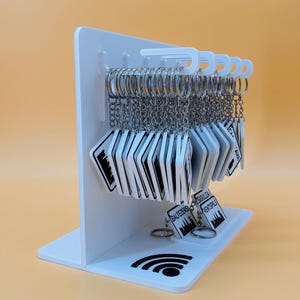 May include: A white acrylic display stand holding multiple square keychains with silver metal rings. Each keychain features a black and white graphic design with text including Baltezers, Sigulda, and Ventspils. A black Wi-Fi symbol is printed on the base.
