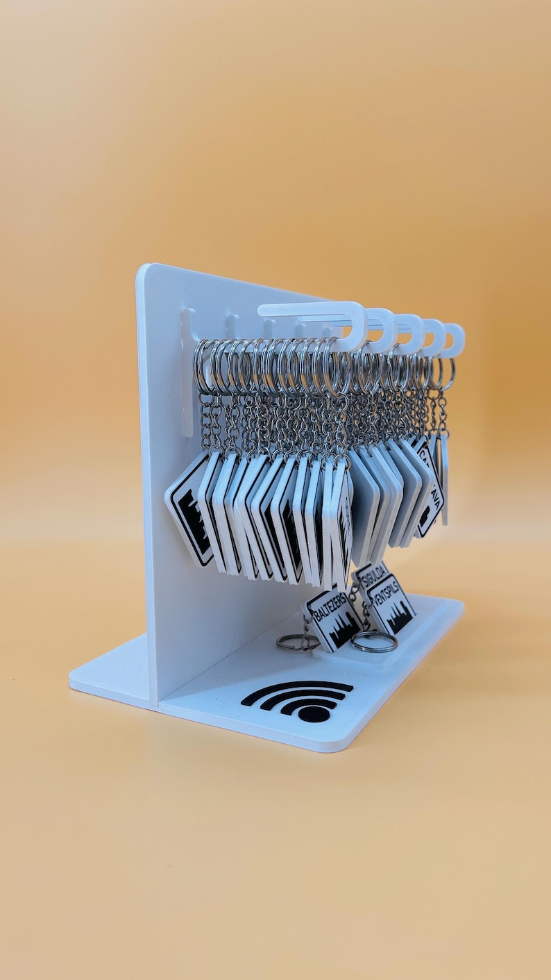 May include: A white acrylic display stand holding multiple square keychains with silver metal rings. Each keychain features a black and white graphic design with text including Baltezers, Sigulda, and Ventspils. A black Wi-Fi symbol is printed on the base.