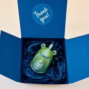 Puede incluir: Un colgante verde con forma de cabeza de oso con una expresión seria, presentado en una caja de regalo azul oscuro. El colgante está sobre un cordón negro, sobre papel de seda negro. Una pegatina redonda dentro de la caja dice "¡Gracias!"
