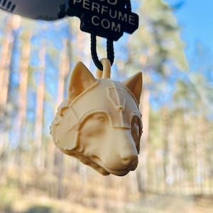 Op de afbeelding: Een beige wolfskop auto luchtverfrisser. De wolf draagt een helm. De luchtverfrisser hangt aan een zwart koord. Een klein zwart label met de tekst "3D PERFUME .COM" is aan het koord bevestigd.