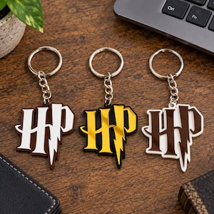 May include: Three metal keychains featuring the letters HP in a stylised font with a lightning bolt design. The first is white with brown edges, the second is gold with black edges, and the third is brown with white edges, each attached to a silver ring.