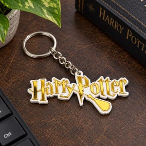 May include: A metal keyring featuring the Harry Potter logo in a shiny gold finish with a white border. The design includes a stylised broomstick integrated into the lettering, attached to a circular silver split ring by a short metal chain.