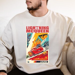 May include: Cream-colored sweatshirt featuring a graphic of Lightning McQueen from the Cars movie. The design includes the text "LIGHTNING McQUEEN" in red, with racing cars and the number "95". The Cars logo is at the bottom.