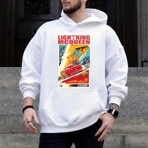 May include: White hoodie featuring a colorful graphic of Lightning McQueen from the movie Cars. The graphic includes the text "LIGHTNING MCQUEEN RACING WORLD" and the number "95". The hoodie has a front pocket and a drawstring hood.