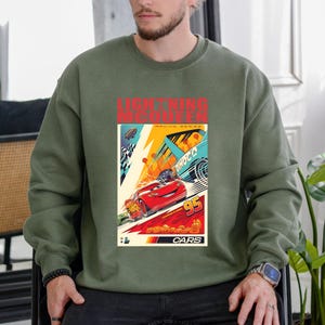 May include: Olive green sweatshirt featuring a graphic of Lightning McQueen from the Cars movie. The graphic includes the text "LIGHTNING MCQUEEN" and "CARS", along with a racing scene with the number 95. The design is in vibrant colors.